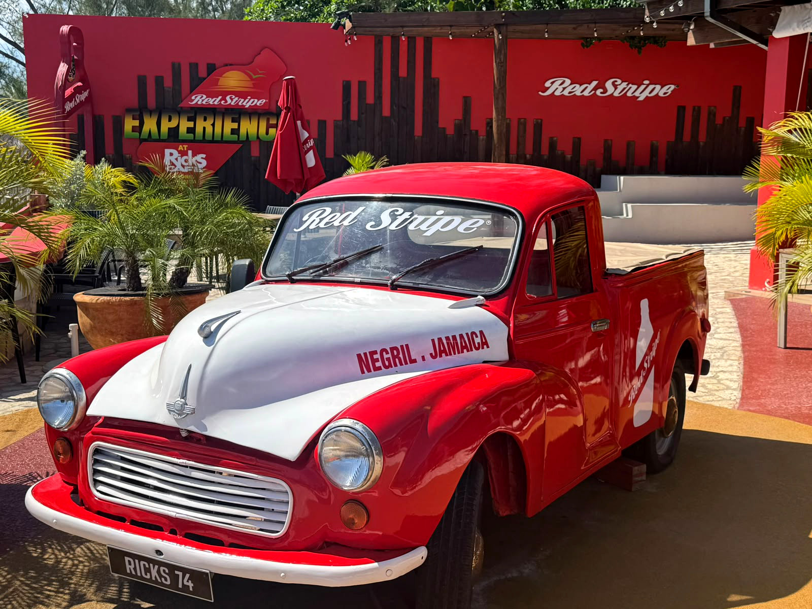 A vintage red‑and‑white Red Stripe pickup truck gleams under the Jamaican sun at Rick’s Café in Negril. Surrounded by tropical plants and vibrant signage, the scene celebrates local culture and adventure — a highlight easily reached through Island Turf Tours’ Negril and Montego Bay airport transfers.