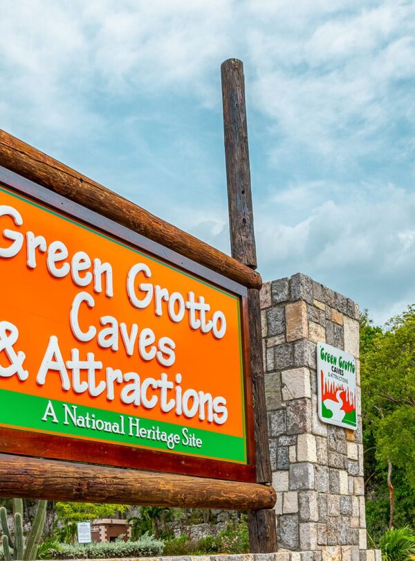 The outside frontal sign of the Green Grotto Caves bears its name as, “Green Grotto Caves & Attractions – A National Heritage Site”, inside a strong wooden framing. A smaller sign with similar wording sits to the right beside it on a cut stone wall. It is a calm evening and many trees and plants litter the background with the very cloudy sky overhead.