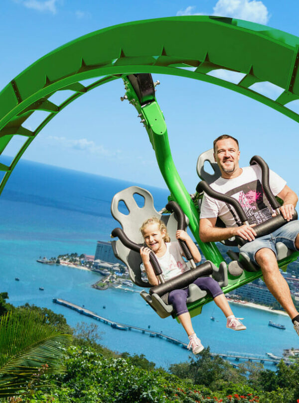 A dad and his daughter in white and painted t-shirts and short jeans experience the thrill of the Raggamuffin Mountain Ride at the Mystic Mountain Adventure Park in Ocho Rios, St. Ann, Jamaica. They hover high above the trees inside the facility on a large green support beam, and in the background the rest of the seaport city can be seen with boats, ships, hotels, residential sections, and mountainsides capping off the experience.