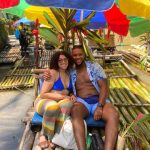 Woman in blue bikini top seated on a bamboo raft beside man with light-blue open shirt and shorts with one arm around her and both smiling with colorful umbrella and other bamboo rafts behind them enjoying their rafting tour.
