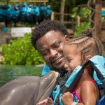 Smiling man introducing excited 4-year old girl to dolphin in water who is close enough for the child to kiss it.