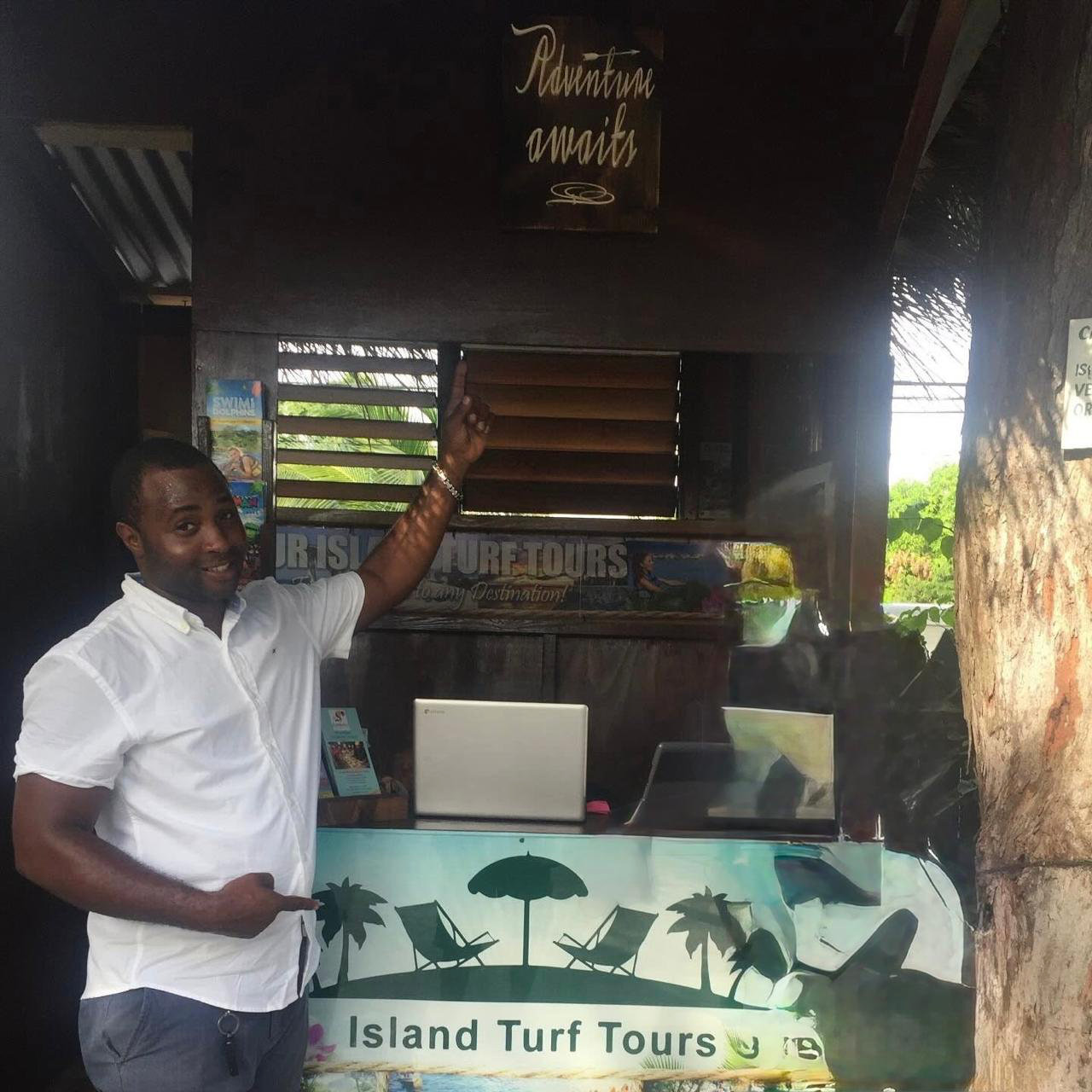 Norris standing to the left at an Island Turf Tours Office Booth pointing up at the slogan, “Adventure Awaits”, with his left hand, and with the other hand pointing down at the Island Turf Tours sign that spans the width of the booth’s table, as if to say, “guys, this is where it’s at”.