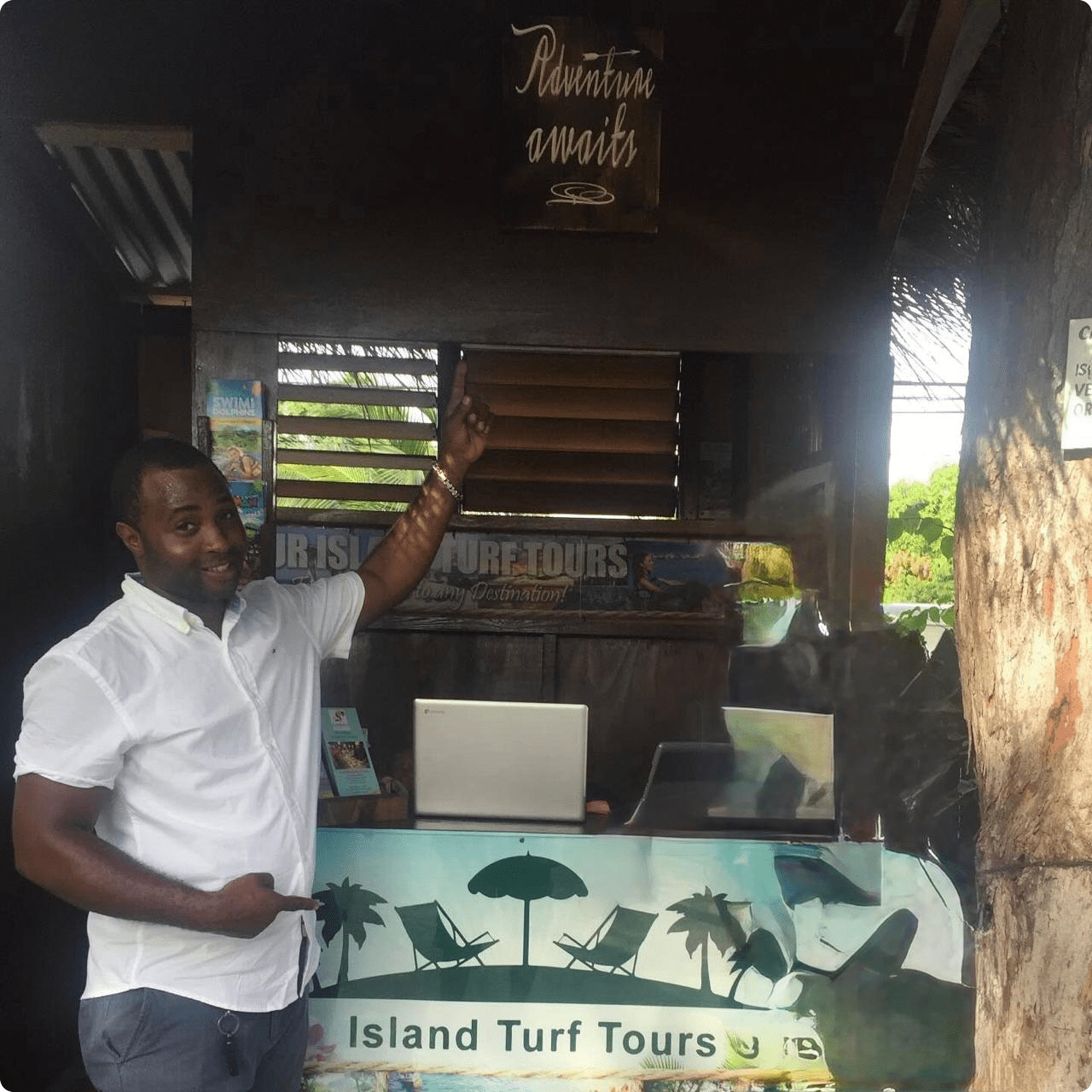 Norris standing to the left at an Island Turf Tours Office Booth pointing up at the slogan, “Adventure Awaits”, with his left hand, and with the other hand pointing down at the Island Turf Tours sign that spans the width of the booth’s table, as if to say, “guys, this is where it’s at”.