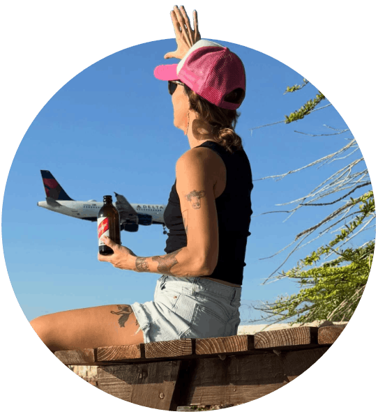 Norris’ private transfers guest sits casually on a bench on beach in Montego Bay in a black merino tank top and short shorts and waves to a Delta Airline plane passing overhead. She is smiling and in high spirits as she has her complementary supplied Jamaican Red Stripe beer in her other hand from Island Turf Tours.