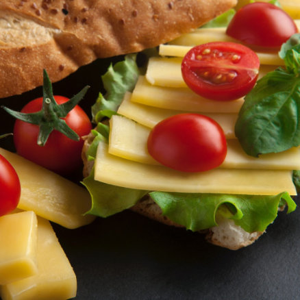A dark colored table that has a close-up shot of sliced cheese, cut tomatoes and lettuce on top of an offset long sandwich fresh bread.
