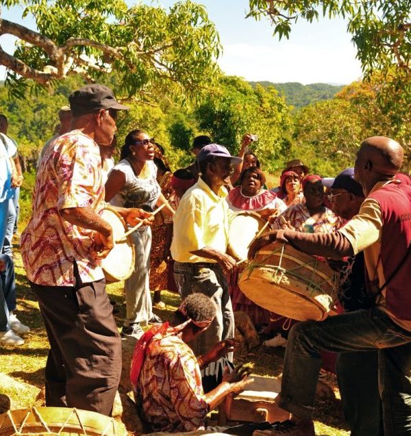 In Accompong Maroon Village, this scene captures a cultural setting of indigenous musicians, some dressed in cultural African garbs and the women wearing ‘Bandanas’, which is an unofficial Jamaican fabric. Some play on drums and other percussion instruments while singing while onlookers are all around on this bright sunny morning. One person raises his digital camera over the scene to take a picture. The hills of the Cockpit Country can be seen in the background. Norris would take his private tour guests here who crave for a taste of our island’s cultural past.