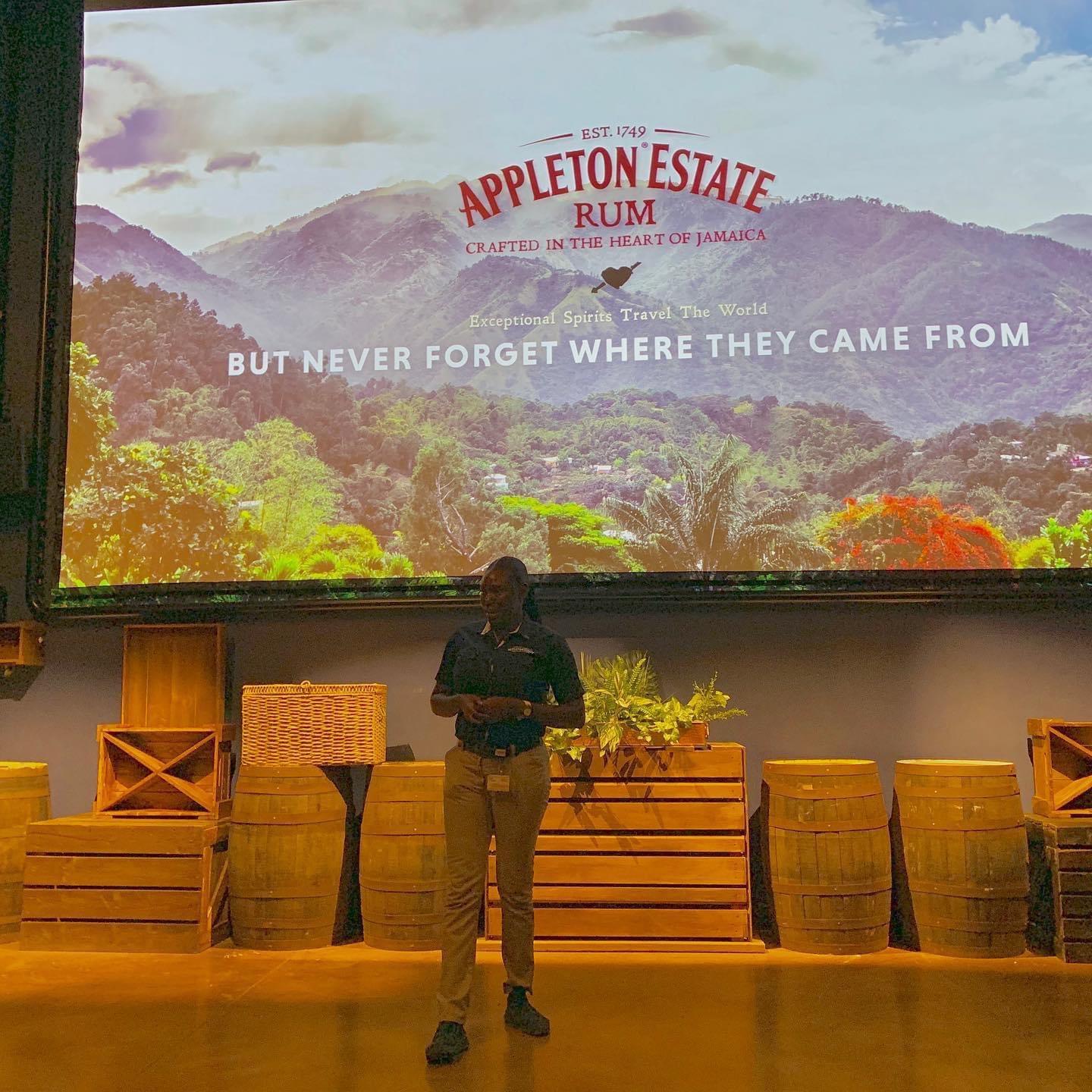 An employee of the Appleton Estate rum tour stands underneath a slide presentation preparing to field questions from a traveling group. On the screen above her is the end of the presentation showing the Appleton logo and beneath it, the words, “Exceptional spirits travel the world, but never forget where they came from”. Located in Santa Cruz, St. Elizabeth, the Appleton Estate Jamaica rum tour is frequented by travelers to Jamaica as well as by local guests of Island Turf Tours.