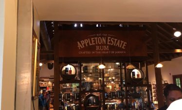 An over-the-shoulder view of a stand that houses several sealed bottles of Appleton rum products. In the foreground to the right is an attendant, half-shown, receiving payment from a guest of the Appleton Rum Tour Experience whose left shoulder sticks into view. On the stand is a heading sign that says, “EST. 1749 Appleton Estate Rum. Crafted in the heart of Jamaica”. In the background are a few other guests strewn about who are looking at other exhibits at the tour. Norris often caters to those who appreciate the laid back nature and history of rum-making in Jamaica when he carries them to this experience in Santa Cruz, St. Elizabeth.