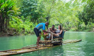 Two Island Turf Tours guests sit intrigued on their raft as their raft driver rubs limestone paste onto the feet of the female passenger, as she has her arm on the other passenger and is smiling. The Lethe River river is calm in the background with lush trees and vegetation surrounding the banks of it, under a calm sunless evening.