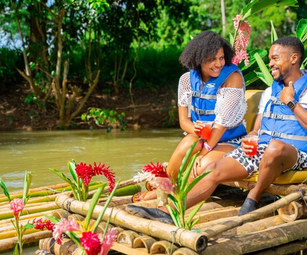 Two female and male guests sit teasing each other, smiling with red drinks in their hands, with the man pointing to himself for conversational effect and the woman looking at him sarcastically. It’s a sunny day on the Lethe River and the bamboo raft they’re sitting on is decorated with the lovely floral Pink Cone Ginger Plant with bright red and pink colors along with their beautiful long green leaves.