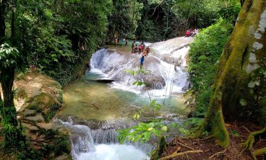 A scene with two distant scattered groups of people enjoying standing on the watery rocks at Benta Falls, a popular tourist destination for Island Turf Tours guests, located in Westmoreland. The bark of a tall tree can be seen to the right of the foreground with lush greenery surrounding the entire area.