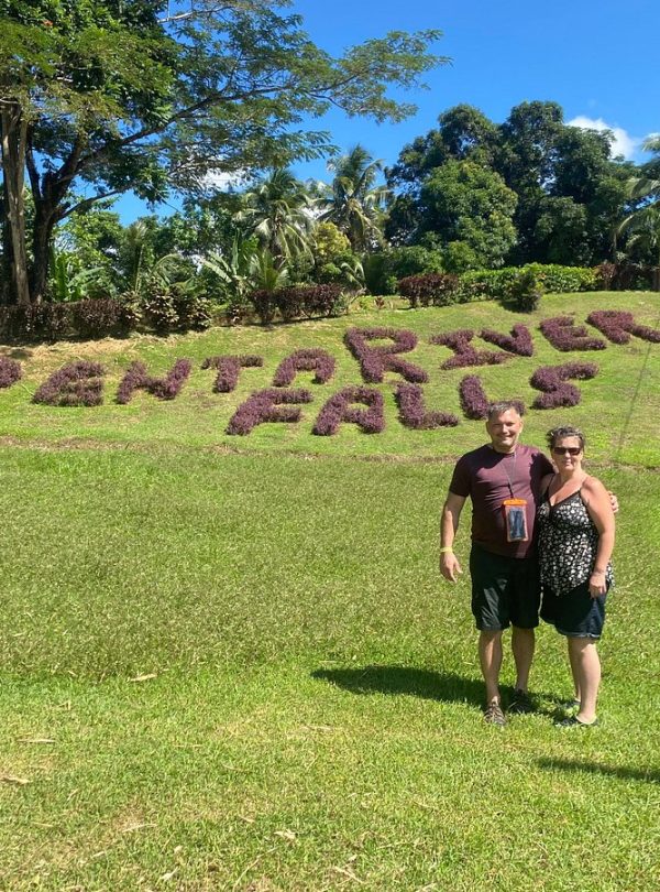 A couple, Norris’ tour guests, stands and smiles at the open area of Benta River Falls where the text “Benta River Falls” can be seen on the floor having been created from tightly grown groups of small purple flowers. The entire scene is filled with lush plant life.