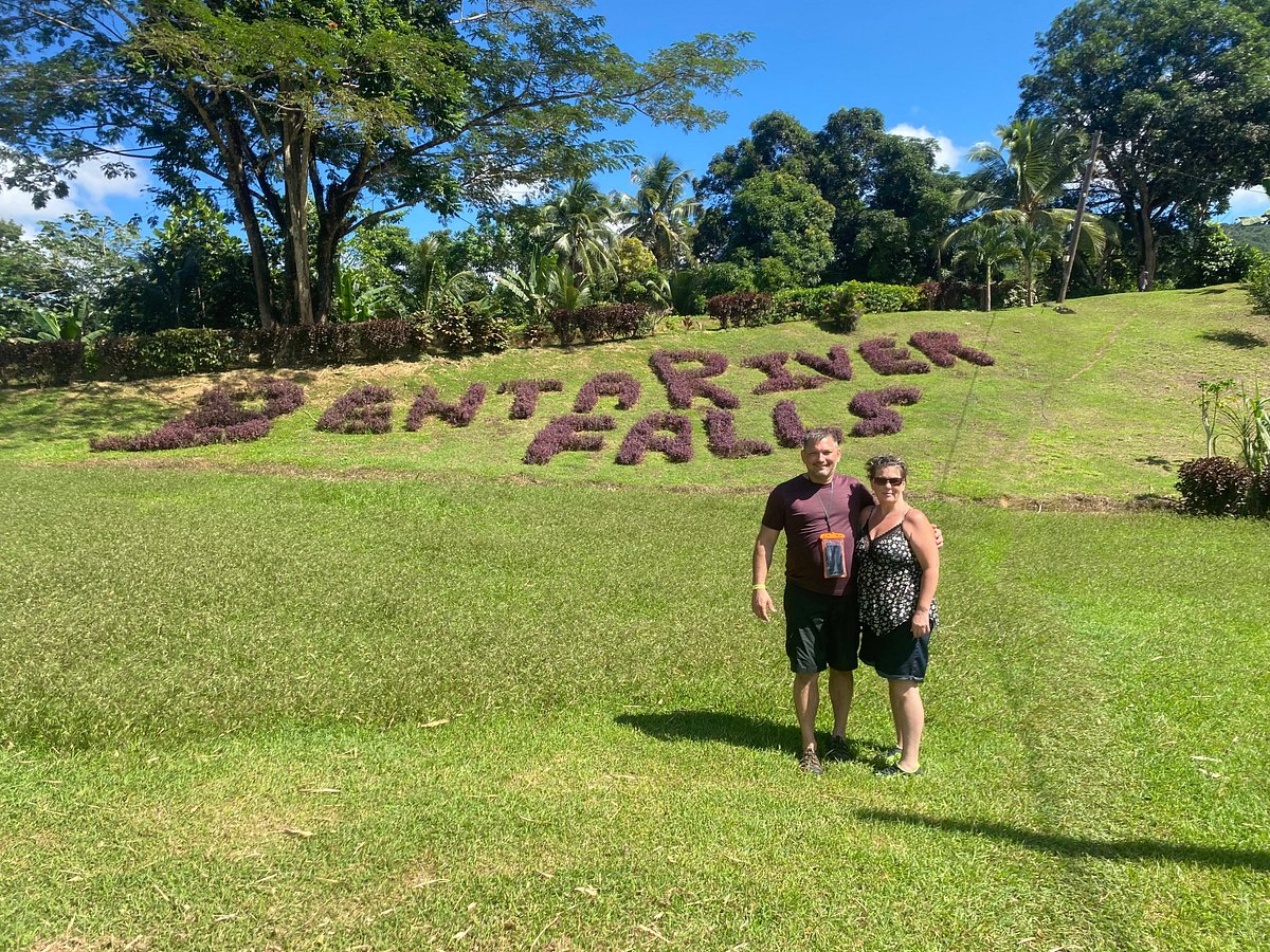 A couple, Norris’ tour guests, stands and smiles at the open area of Benta River Falls where the text “Benta River Falls” can be seen on the floor having been created from tightly grown groups of small purple flowers. The entire scene is filled with lush plant life.