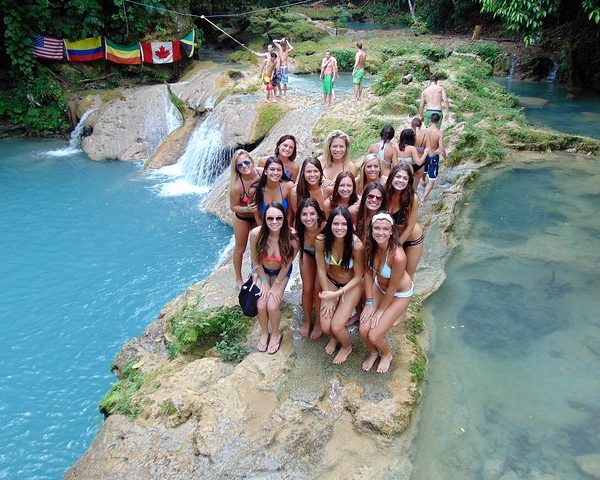 In the cool of the evening, we see a lower walking platform section to the taller, watery falls of the Blue Hole in Ocho Rios sees 13 lovely young ladies pose for a picture to be taken of their Island Turf Tours trip to the swimming attraction. 10 others are off behind them in the distance, 6 of them being male. On the upper left of the shot are some flags spread out along the jungle plants overlooking the waters: one each representing the USA, Columbia, Ethiopia, Canada, and Jamaica.