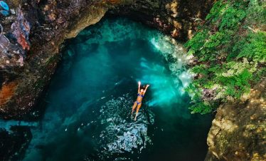A top-down view inside the Blue Hole, of one of Norris’ guests in a blue bikini floating on her back on the water top in a picturesque scene with the rocks and the shrubs as the backdrop.
