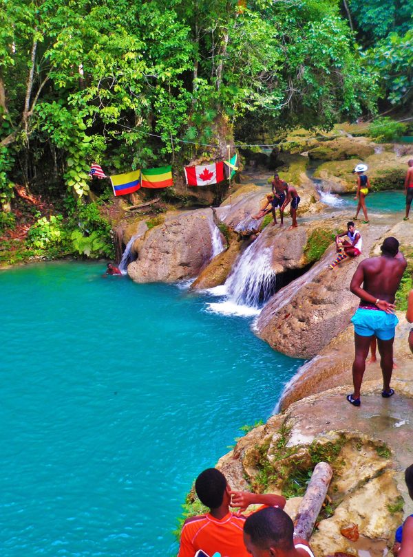 As the evening winds down at the lower walking platform at the Blue Hole in Ocho Rios, St. Ann, many travelers to the swimming attraction are walking around and admiring its surroundings. Of the 28 persons present, some are swimming in the blue-green waters, some are talking before they jump off again into the natural pool, and some are just looking about. Five flags can be seen in the distance spread out and hanging on the sides of the wooded area, representing the USA, Columbia, Ethiopia, Canada, and Jamaica. The atmosphere is one of lush greenery and beautiful watery basins. Some were carried here by Norris of Island Turf Tours.