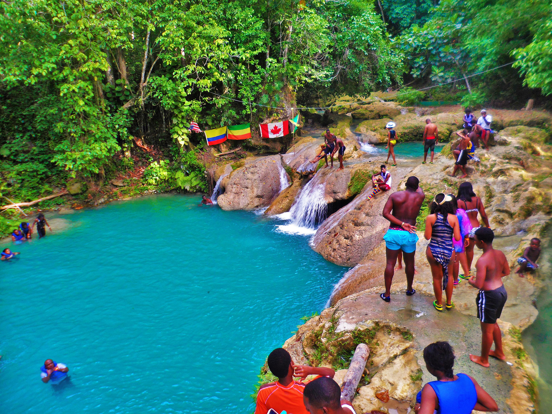 As the evening winds down at the lower walking platform at the Blue Hole in Ocho Rios, St. Ann, many travelers to the swimming attraction are walking around and admiring its surroundings. Of the 28 persons present, some are swimming in the blue-green waters, some are talking before they jump off again into the natural pool, and some are just looking about. Five flags can be seen in the distance spread out and hanging on the sides of the wooded area, representing the USA, Columbia, Ethiopia, Canada, and Jamaica. The atmosphere is one of lush greenery and beautiful watery basins. Some were carried here by Norris of Island Turf Tours.