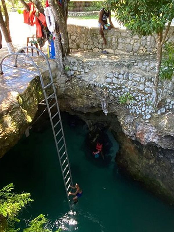 A guest in a blue full-suit bikini climbs down into a section of the Blue Hole using the provided ladder, as onlookers at the top watch her go down. At the top are three persons looking in and another seated on a wall with a piece of wood in his hands. A couple is seen already swimming in the water below as well.