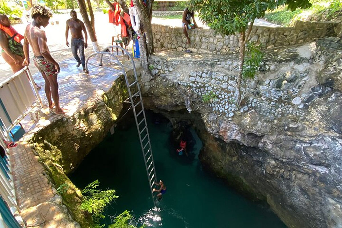 A guest in a blue full-suit bikini climbs down into a section of the Blue Hole using the provided ladder, as onlookers at the top watch her go down. At the top are three persons looking in and another seated on a wall with a piece of wood in his hands. A couple is seen already swimming in the water below as well.