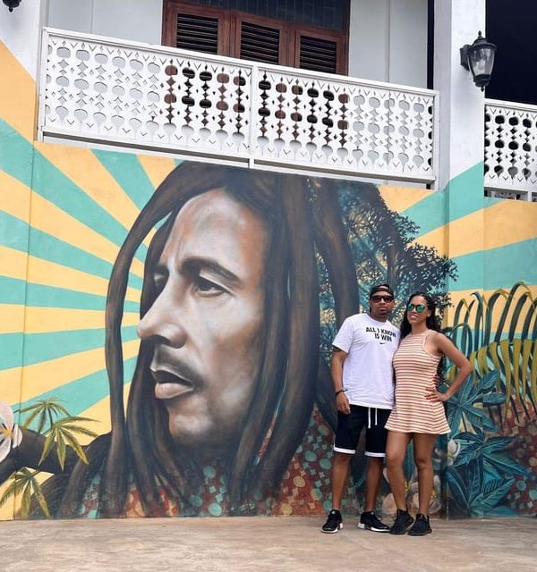 A man and woman couple, guests of Island Turf Tours’ 9 Miles tour, pose with arms around each other before the side of a building that has a mural of Bob Marley’s face looking away, imprinted on the side of it. They are dressed casually and wearing black sneakers and sunglasses – the man in a Nike shirt and the woman in a short striped dress.
