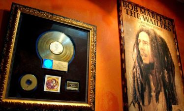 Two pictures on an orange wall with Bob Marley on the right, with the words over his head saying, “Bob Marley and the Wailers”, and the picture on the left showing a large golden record atop a golden CD and a golden Cassette, with an inscription under the record, saying “Presented to Bob Marley”, for the record label “Confrontation”.