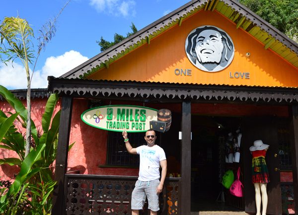 One of Norris’ 9-Mile tour guest smiles and stands in front of a colorful building that has a woodcarving of Bob Marley and the words, “One Love” on the inset of the triangular roof, and he points right up to an oval wooden sign that says, “9 Miles Trading Post”.