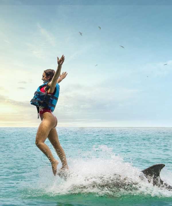 A woman stands on one leg, arms outstretched, balanced on the nose of a dolphin as he rides with her on a promo shot for the Dolphin Bay tour located at Montego Bay and Negril in Lucea, Jamaica. The Caribbean Sea is in the background. Private tour traveling guests of Island Turf Tours at times ask to come to this attraction.
