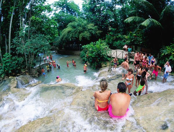 A view from the low height of a rocky mound of the world famous Dunn’s River Falls, in Ocho Rios, St. Ann, Jamaica. Two guests are seated one part of the rock in the foreground while many others are scattered all throughout the area. This is a popular evening scene showing just how many tourists flock to the Falls to frolic and enjoy themselves in the beautiful natural scenery.