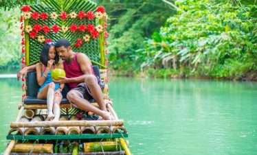 Two of Norris’ tour guests couple up snuggly on a nicely decorated raft as they drink water from a coconut that the man is holding up. The scenery on the Martha Brae marsh is quiet and the water is calm, and everywhere is green round about them with overhanging tree limbs, and the raft is dressed in a red and yellow sequence of Hibiscus flowers and a woven set of palm branches at the back of the raft.