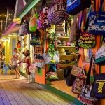 A brightly yellow-lit shot of the Montego Bay Craft Market shows a lot of indigenous craft items and souvenirs, like bags, hats and caps, and other items, most of which has the word, “Jamaica” written on them. A little girl and boy can be seen to the left exiting one of the vendor’s store to rejoin the small group that just had an airport transfer from Island Turf Tours.