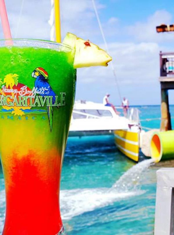 A glass of mixed-colored drink is prominently focused on to the left of this scene, with green as the top drink mixture, yellow in the center and red at the base, and the words, “Jimmy Buffett’s Margaritaville Jamaica” in the logo shown. Two straws peek out the top and a piece of pine and with a cherry and toothpick stuck in it hangs off to the right. Margaritaville, Montego Bay is a popular destination for Island Turf Tours guests.