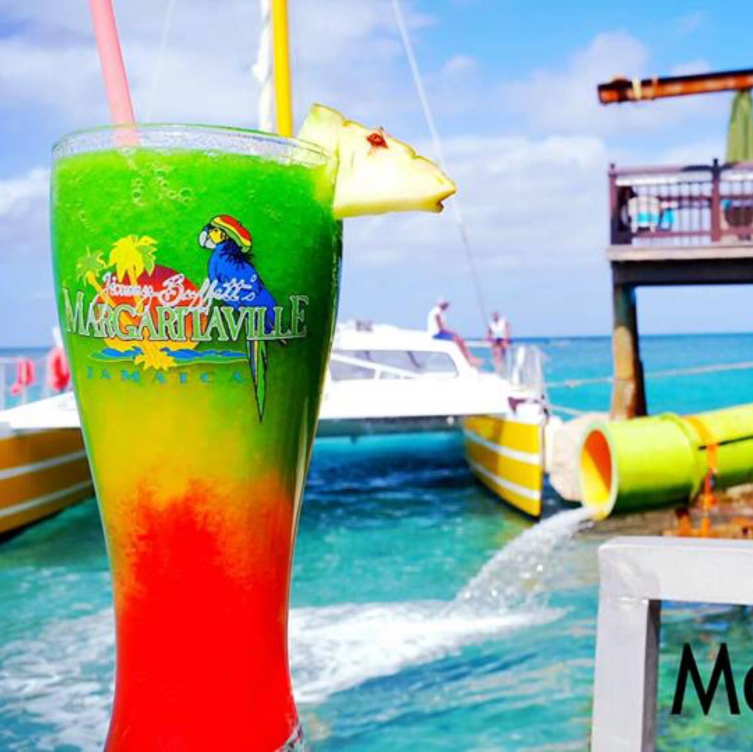 A glass of mixed-colored drink is prominently focused on to the left of this scene, with green as the top drink mixture, yellow in the center and red at the base, and the words, “Jimmy Buffett’s Margaritaville Jamaica” in the logo shown. Two straws peek out the top and a piece of pine and with a cherry and toothpick stuck in it hangs off to the right. Margaritaville, Montego Bay is a popular destination for Island Turf Tours guests.