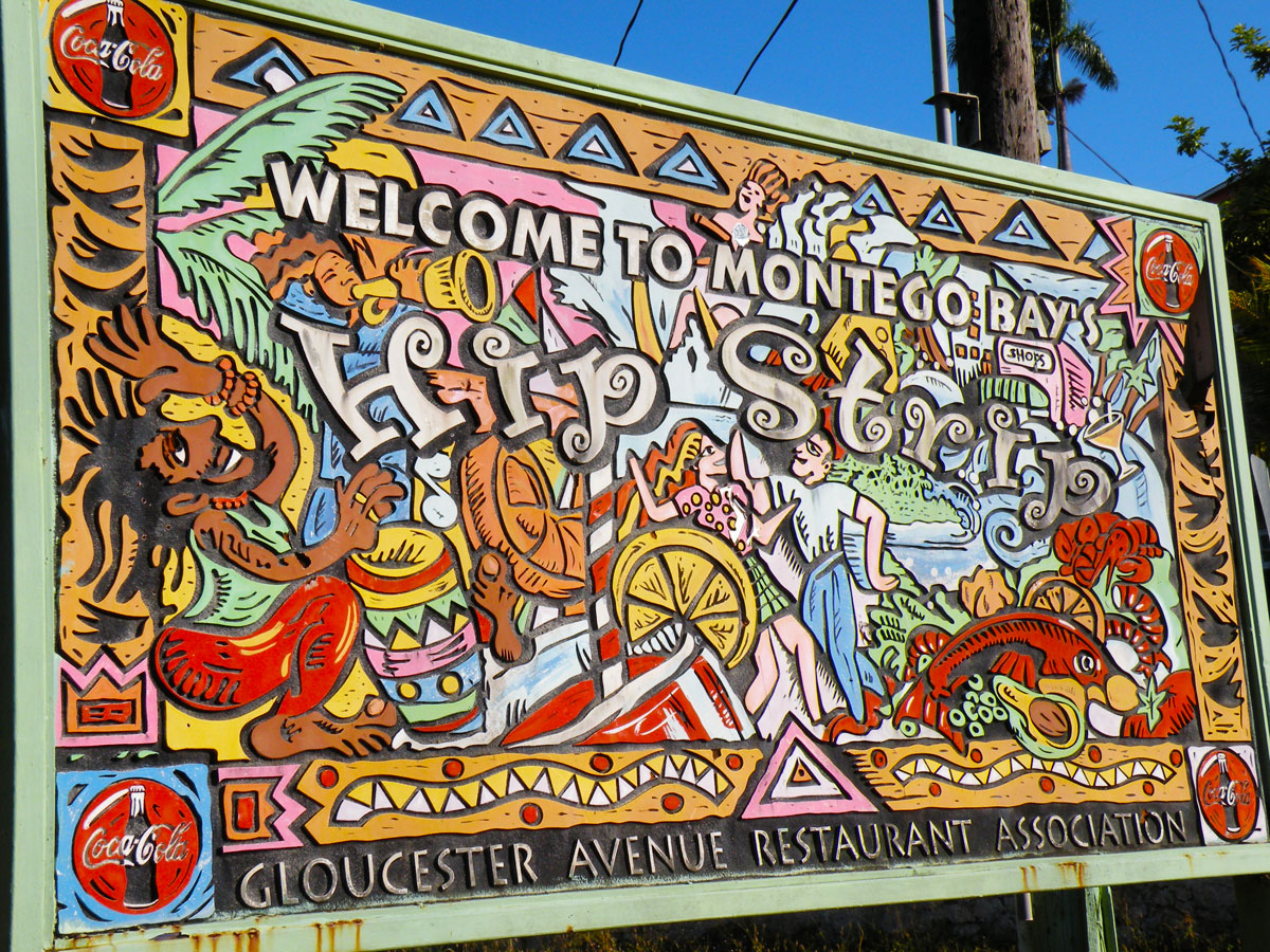 The main 2+ meter sign of a colorful wooden-created mural relief welcomes guests to the city by saying “Welcome to Montego Bay’s Hip Strip”, put up by the Gloucester Avenue Restaurant Association. It shows a Rastafarian playing the drum on the left, a flute-player at the upper left some drinks in the foreground, a couple dancing and having a great time, and food and shops on the right.