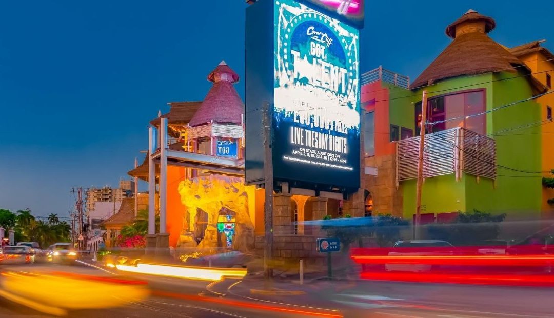 The colorful streets of Montego Bay light up in the evening as cars whizz by in the popular city for Norris’ guests. A Large Media screen sign showing “Coral Cliff Got Talent” is prominently seen showing an advert for the local talent auditions, and a small Burger King sign sits calmly beneath it. Palm trees can be seen off in the distance on the opposite left.