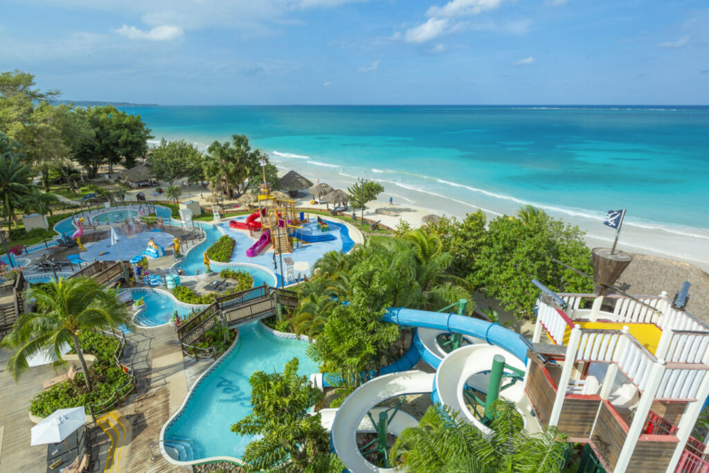Colorful Negril-based resort, Beaches hotel brand location showing vast interlocking pools section with children’s water slides, palm trees, and other tropical trees interspersed, and with the beach waters off in the background.