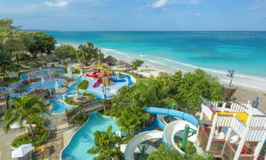 Colorful Negril-based resort, Beaches hotel brand location showing vast interlocking pools section with children’s water slides, palm trees, and other tropical trees interspersed, and with the beach waters off in the background.