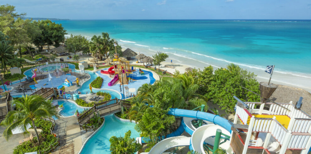 Colorful Negril-based resort, Beaches hotel brand location showing vast interlocking pools section with children’s water slides, palm trees, and other tropical trees interspersed, and with the beach waters off in the background.