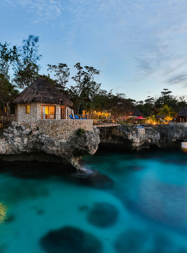 The Rockhouse Hotel in Negril is a popular spot for carrying visitors to Jamaica via Island Turf Tours airport transfers and hotel transfers. Here in an evening scene the lights turn on inside the many stone and rocky hotel hut-like villas that sit lining the land’s edge overlooking the Caribbean Sea. A famous one-sided jumping bridge is seen in the center where patrons can jump off from, land in the water, and then swim a few meters to climb back up on a ladder that reaches down into the sea.