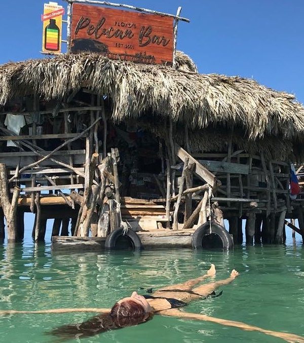 A lone tourist guest of Island Turf Tours can be seen here in the foreground right outside Floyd’s Pelican Bar in a close-up shot floating on her back in her two-piece black bikini. It would appear like she’s relaxing in water that seems to stretch out for miles beyond the wooden bar structure. On the top of the ‘floating’ building is a two-sided sign that has a Red Stripe beer picture hanging off to the left of it, and on the right it says, “Floyd’s Pelican Bar. Est. 2001”.