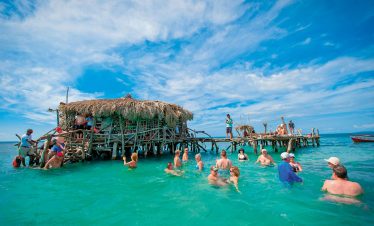 A sunny but still cool (not pelting sun) outdoor shot showing many guests of the Pelican Bar hanging out together inside the bar, some seated, some standing, hanging out on the walkway of the bar, and in the foreground, persons inside the waist-height water outside in the green watery sea. Many adults, some of the men wearing caps, and the women in their swim-suits, hold their bottles of Red Stripe Beer while they’re enjoying their time being in the water and having their enjoyable conversations. A small fishing-type boat sits off to the right with the words, “American Queen” written on its side, and in the far background the side of a mountain edges out into the sea behind it. For travelers visiting Jamaica Floyd’s Pelican Bar, roughly one mile offshore from the Parottee Bay in the southwest St. Elizabeth parish of the island, is an interesting sight that attracts tourists, some of whom seen here are guests of Island Turf Tours who enjoy this Jamaica private tours transport service.