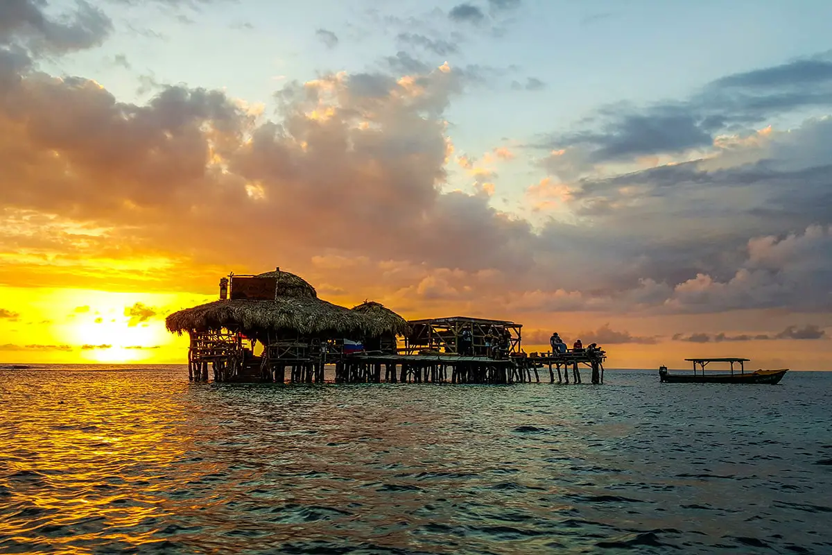 An evening scene of Floyd’s Pelican Bar showing the sun going down just to the left of the hut-like bar standing on stilts and with the entire Caribbean Sea behind it. There are a few Montego Bay to Negril airport transfer Island Turf Tour guests sitting on the walkway and some standing and having conversations too. A small fishing boat that carried them to the spot sits in the evening silhouette off to the right.