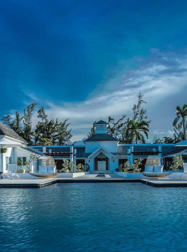 A dull-blue dusk scene inside the pool section of the Trident Hotel in Port Antonio, Portland, Jamaica. In the foreground is the water of the pool and surrounding it are several white umbrellas with pool lounge chairs underneath. Other canopy seats can be seen and the facility is in the backdrop with many trees in the surroundings.