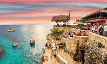 Visitors of Ricks Café overlook the balcony at three becalmed boats in the blue-teal ocean, and breathtaking iridescent sky. In the foreground, a pergola serves as refuge from the incandescent rays of the sun. The rockface steps fully illuminated by the sun's rays, descends from the cafe to the shoreline serves as additional vantage points observe the beauty of the ocean and sky.