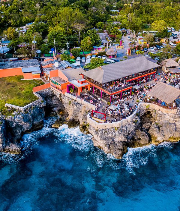 Aerial view of Rick’s Café overlooking the massive boulder rockface of the shoreline. Dense foliage in the background contrast with the brilliant blue of the ocean smashing against the coastal cliff below the restaurant. Crowds of visitors dot customer spaces. At the foremost point, surrounded by gazebos, and three cabanas below a red Rick’s Café 1974 sign, a pool houses additional groups of guests.