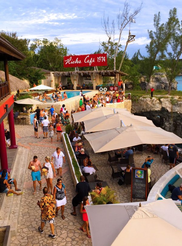 Captured from the second balcony of the Rick’s Café, an aerial view of the customer spaces littered with tan fabric shade sails. Guests with relaxed postures amble around the cobblestone area. In the immediate foreground, one fabric shade partially visible with three more overlooking the scene of one of Island Turf Tours’ most popular stops.