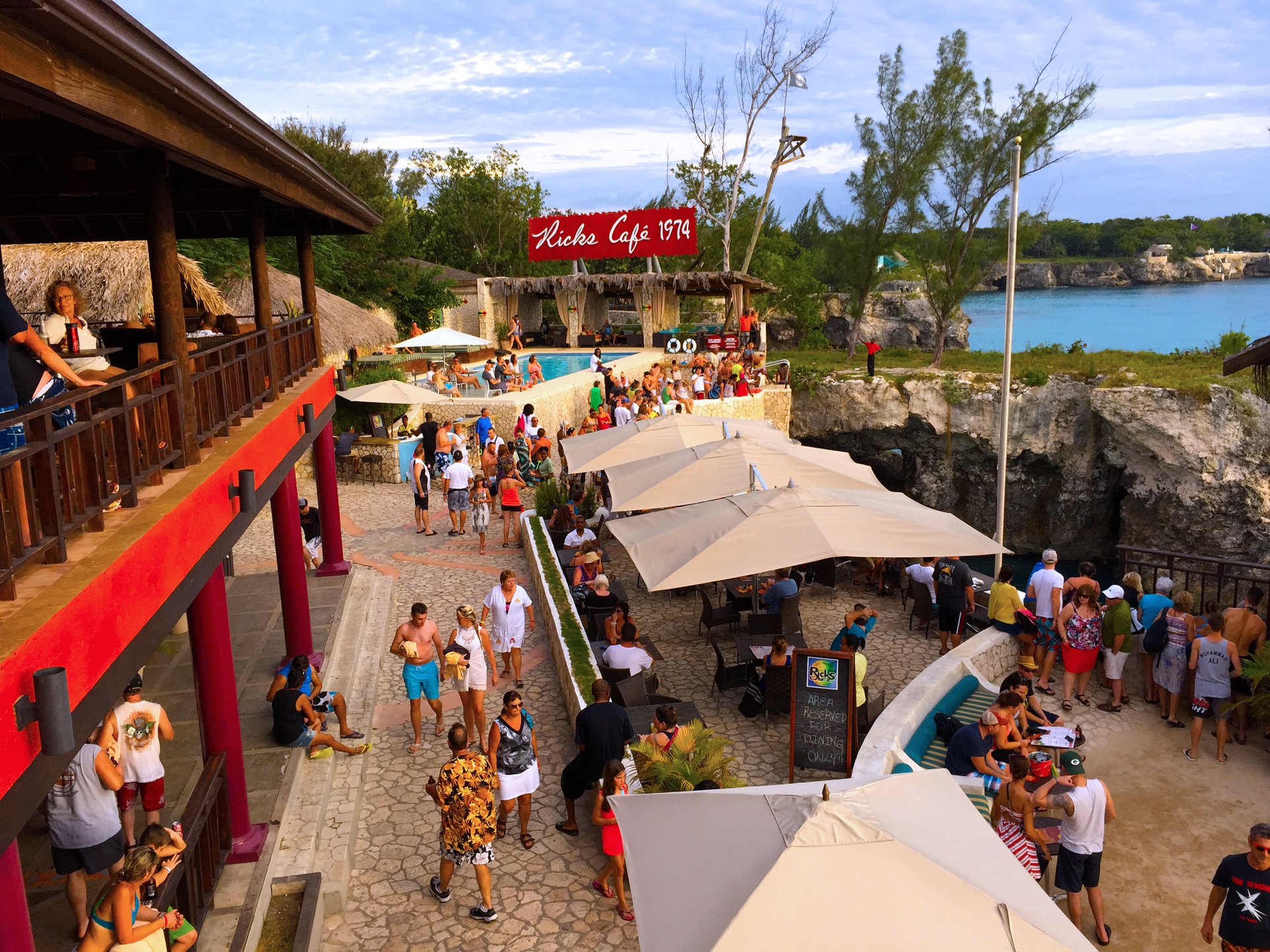 Captured from the second balcony of the Rick’s Café, an aerial view of the customer spaces littered with tan fabric shade sails. Guests with relaxed postures amble around the cobblestone area. In the immediate foreground, one fabric shade partially visible with three more overlooking the scene of one of Island Turf Tours’ most popular stops.