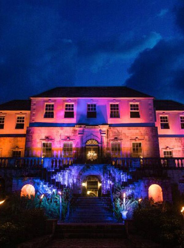 A night time shot of the Rose Hall Great House in Montego Bay, Jamaica, showing how the estate looks during their ‘haunted night tours’. The sky is dark blue and the property is lit up in purple and blue and varying colors. The grounds also have bonfires lit at the front and the chandeliers inside the main are turned on.