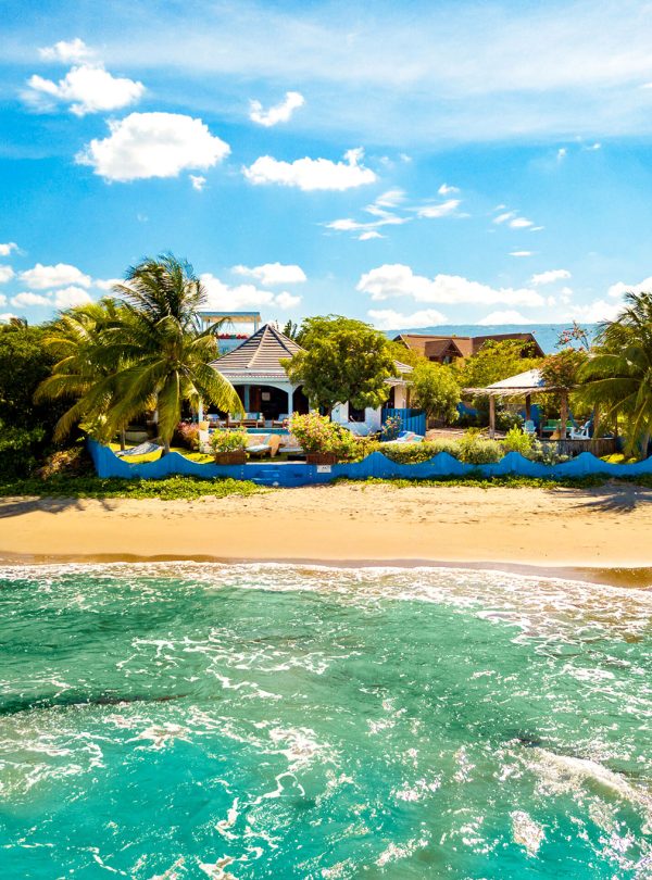 Travelers to St. Elizabeth, Jamaica enjoy themselves when they are carried by Norris to Treasure Beach. Today, in a slightly elevated view from the sea, a relatively calm sea water brushes up against the shore that’s immediately in front of an eating and dining area painted in light blue, and with bright blue short outer walls. A hammock is the left of the enclosed property and other lawn chairs are strewn about. Other businesses and residential areas can be seen in the background with their rooftops just peeking above the lush greenery.