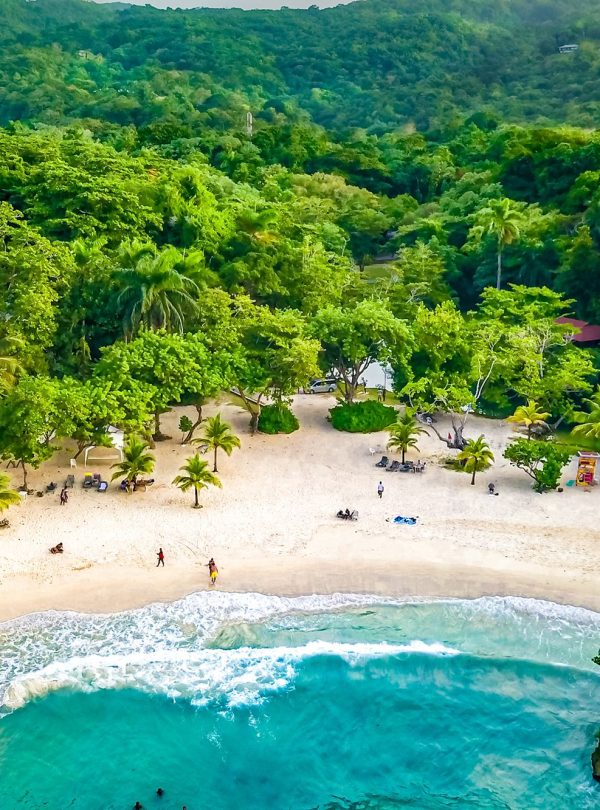 An aerial shot from just above the sea shore of the iconic small, arch-shaped enclosed beach area of Frenchman’s Cove in Port Antonio, Portland, Jamaica, showing an impressively beautiful view of the rough beach water rushing up to the clean sandy shore. About 18 guests to the beach, a few of whom were carried by Island Turf Tours. There are small palm trees that dot the sandy section along with so many other plants. A couple of eating area structures are on the left and right of the scene and the whole property is engulfed in forested trees.