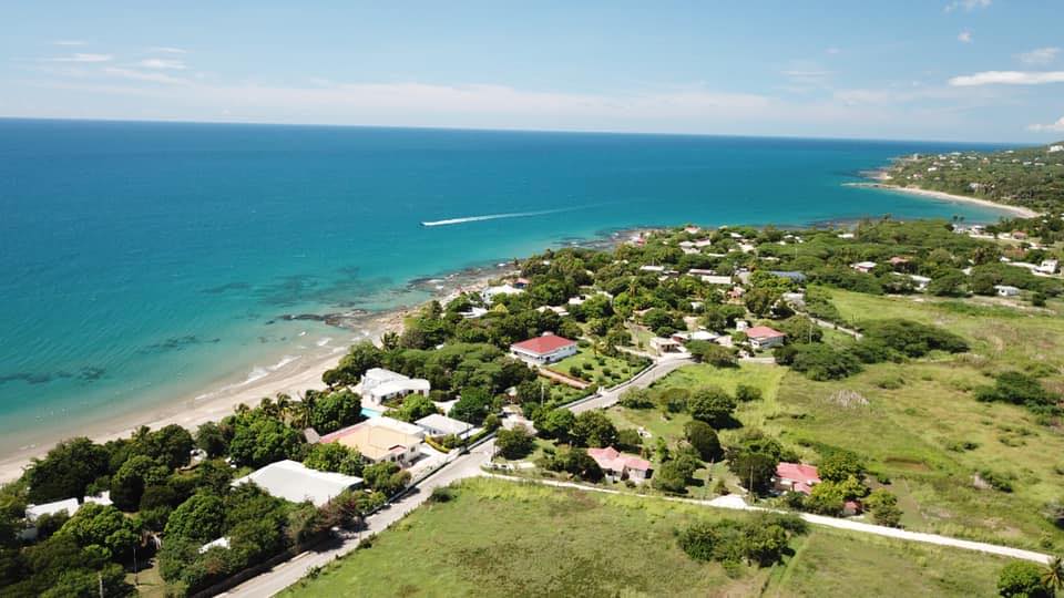 A bird’s eye view of the Treasure Beach in St. Elizabeth parish of Jamaica shows a stretch of the beach flanked by business places that serve the beach, houses and the inward land and roadway to the right. On a bright and sunny day only the streak of water emitted from a lone personal watercraft is seen in the upper center area coming down toward the left of the scene. On the right beyond the houses with their thick trees are grassy pasture areas with a few trees here and there.